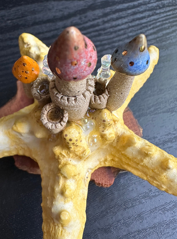 Ceramic sculpture of a yellow sea star with a sand colored castle atop of it, with multicolor pointed tower roofs. The sculpture sits atop a natural piece of wood.