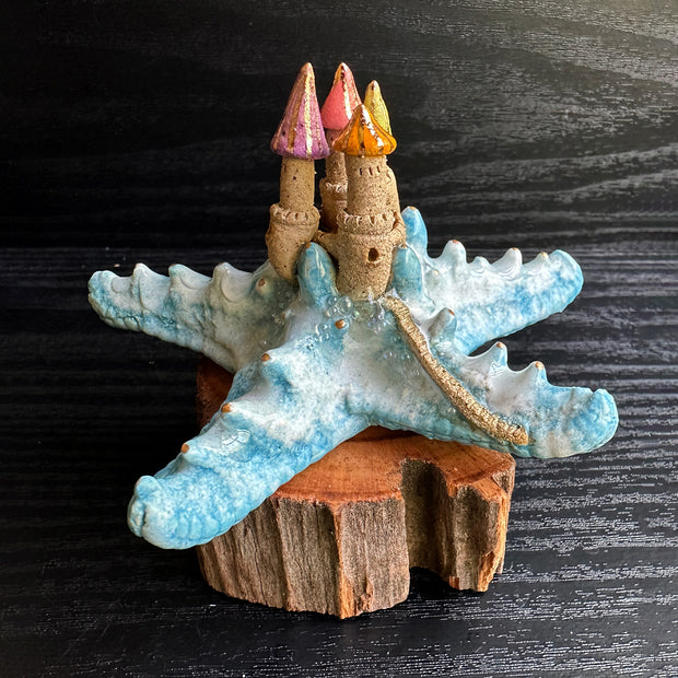 Ceramic sculpture of a blue sea star with a sand colored castle atop of it, with multicolor pointed tower roofs. The sculpture sits atop a natural piece of wood.