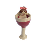 Ceramic sculpture of a crying girl with a pink hair bow and ponytail. She sits inside of a wine glass.