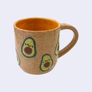 Ceramic mug with spotted finishing and an earthy brown exterior and orange interior. On the outside are painted on cartoon style avocados, with simple expressions.