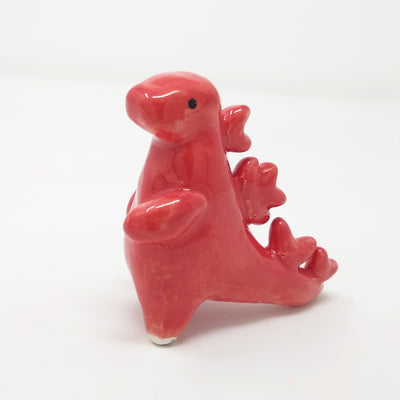 Ceramic sculpture of Godzilla, made of smooth shapes and features. It has stylized spikes on its back and a set of rendered eyes on its otherwise plain face. Colors are glossy reddish pink.