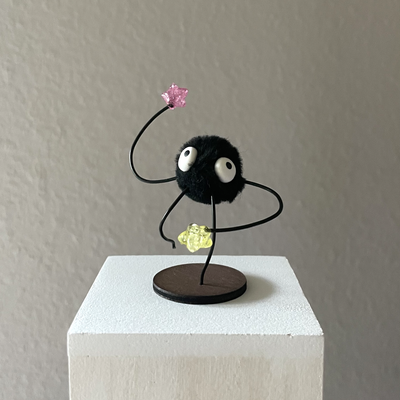 Sculpture of a small black soot sprite, made of a pom pom. It has thin wiry arms and legs and holds 2 semi clear stars.