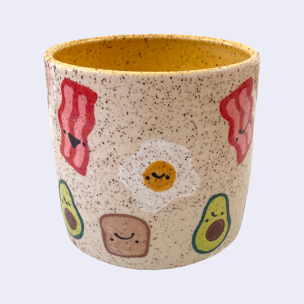 Ceramic planter with spotted finishing and an earthy cream exterior and yellow interior. On the outside are painted on cartoon style eggs, avocado, toast and bacon, with simple expressions.