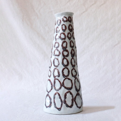 White ceramic vase that narrows at the top, with melted looking brown circles all over it as a pattern.