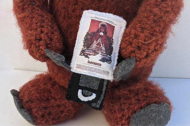 Large crocheted sculpture of Ludo from Labyrinth. He has messy hair and holds a copy of Labyrinth on VHS, which pops out of its case to reveal a crocheted disk.