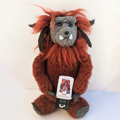 Large crocheted sculpture of Ludo from Labyrinth. He has messy hair and holds a copy of Labyrinth on VHS, which pops out of its case to reveal a crocheted disk.