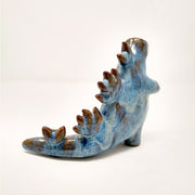 Ceramic sculpture of a Godzilla monster, with an overall smooth body shape and no distinct facial features. It has spikes running down its back and has one hand extended out slightly. Colors are blue with brown drips.