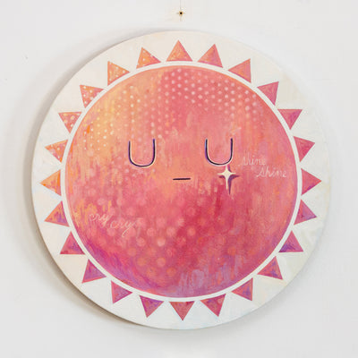Painting of a pink and orange sun with graphic style triangle rays around it. It has a simple, closed eye expression with a gold sparkle on its right cheek. "Cry cry shine shine" is written subtly on its face.