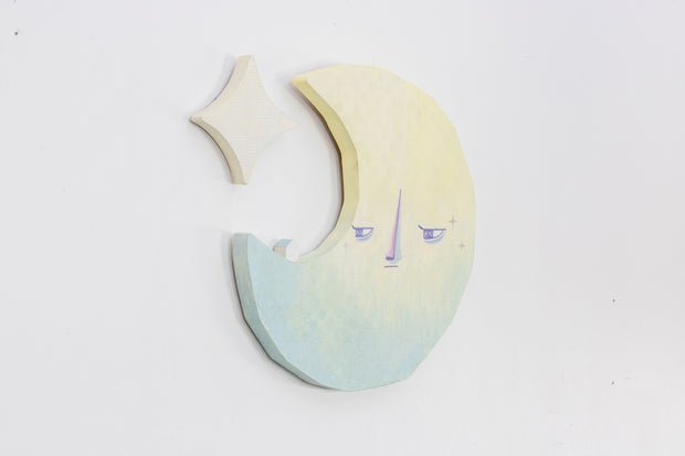 Die cut canvas sculpture of a crescent moon, with an apathetic expression. It looks off to the side at a tiny house that sits on the moon. A star hangs in the upper left corner.