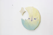 Die cut canvas sculpture of a crescent moon, with an apathetic expression. It looks off to the side at a tiny house that sits on the moon. A star hangs in the upper left corner.