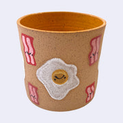 Ceramic planter with spotted finishing and an earthy brown exterior and orange interior. On the outside are painted on cartoon style eggs and bacon, with simple expressions.