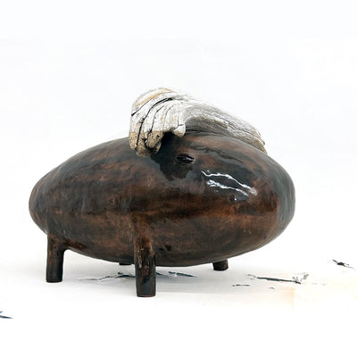 Ceramic sculpture of a large dark brown creature with simplistic shapes and 4 small legs. It has a wood mounted atop its head.