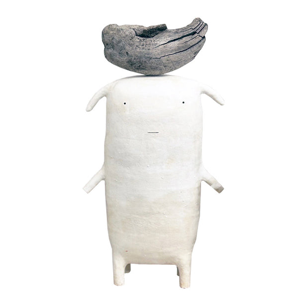 White ceramic sculpture of a simplistic and flat character with short arms and legs and a piece of wood balanced atop its head.