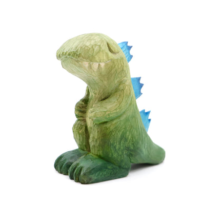 Whittled wooden sculpture of a green Godzilla with blue back spikes, modeled in a non aggressive, cute fashion. Its eyes are peacefully closed and its hands rest on its chest.  