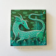 Painting on wood panel of a green dragon with cute simplistic features and winged ears. Its body has scales and it slightly curls into itself, standing in a forest with many small pine trees.