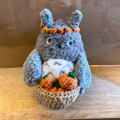 Fluffy crocheted plush of Totoro, with an autumn leaf crown around his head. He holds a brown basket filled with mini pumpkins.