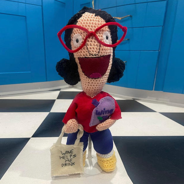Crochet sculpture of Linda Belcher from Bob's Burgers, with a wide smile and holding bagged wine and a tote that reads "Wine Helps Me Drink" on it.