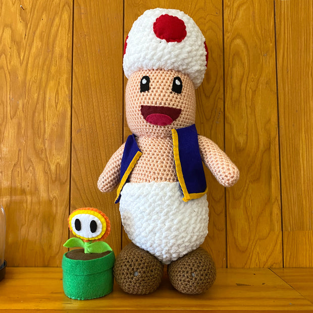 Crochet plush sculpture of Toad from Super Mario Bros., smiling and wearing his usual attire of a vest and white pants. Next to him is a small red and yellow plant from the series in a green pot.