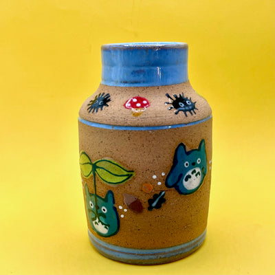 Stoneware vase featuring small, cute illustrations of chibi blue Totoros, holding leaf sprouts with acorns, mushrooms and leaves nearby. Small dust sprites go around the top of the blue lined vase.