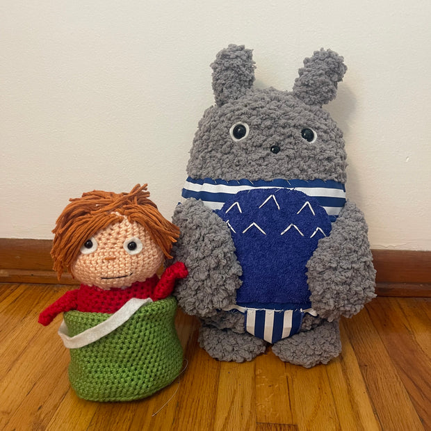 2 crocheted plush dolls, one is of Totoro wearing a blue and white one piece bathing suit. The other is of Ponyo with yarn hair, sitting inside of a green knit bucket.