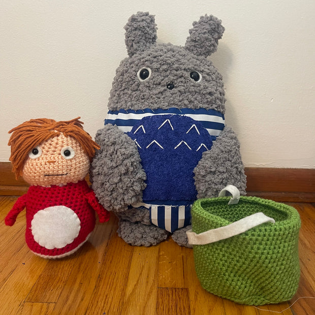 2 crocheted plush dolls, one is of Totoro wearing a blue and white one piece bathing suit. The other is of Ponyo with yarn hair, sitting next to a green knit bucket.