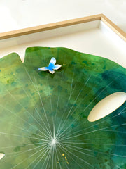 Painting on cut out paper of a round blue and green marble patterned lily pad and thin white lines. A small blue and white butterfly sits atop the pad. Displayed at an angle to show sheen of the resin coating.
