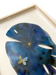 Painted cut out lily pad leaf coated in resin, dark blue with subtle galactic marbling pattern and thin gold stripes. A small blue and gold butterfly rests atop the leaf. Displayed at an angle to show sheen of resin.
