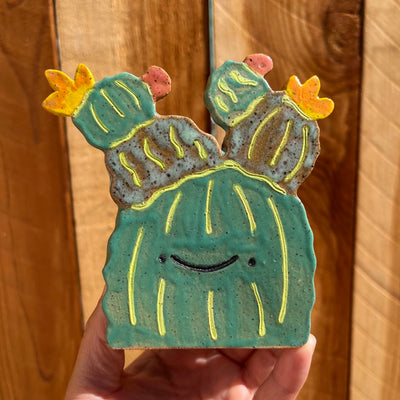 Flat ceramic sculpture of a cactus with barrel like appendages atop its head. It is simplistic in form and has a smiling cartoon face. It has several flowers blooming atop of it.