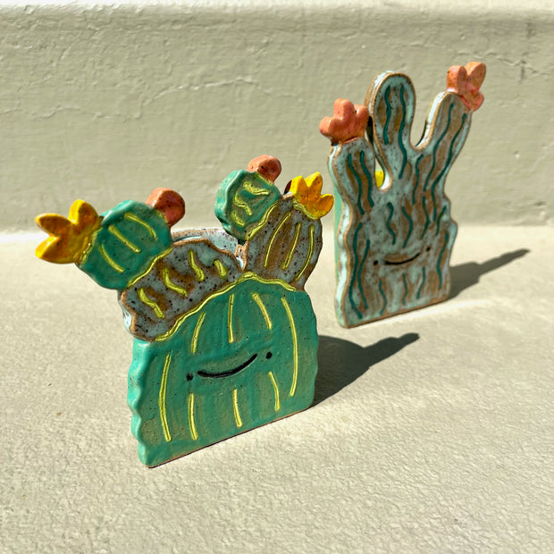 Ceramic sculpture of 2 flat cartoon style cacti, both with smiling faces and simplistic features. Each have flowers blooming from them.