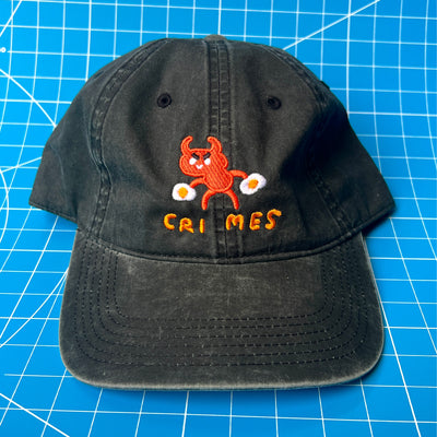 Black cap with a small embroidery in the center of a cartoon red devil with a sunny side up egg in each hand. Below it reads "crimes" which a space between "cri" and "mes."