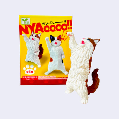 Small fluffy cat figure, standing on its hind legs with one arm positioned up in a power pose, a yelling expression to match. It stands next to its product packaging. 