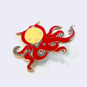 Die cut enamel pin designed by James Jean, of a red octopus with sparse blue and yellow swirl patterning, and a circular human like face, looking down. Its many arms are curled into itself and have blue tentacles. Shown at the side to display dimensionality. 