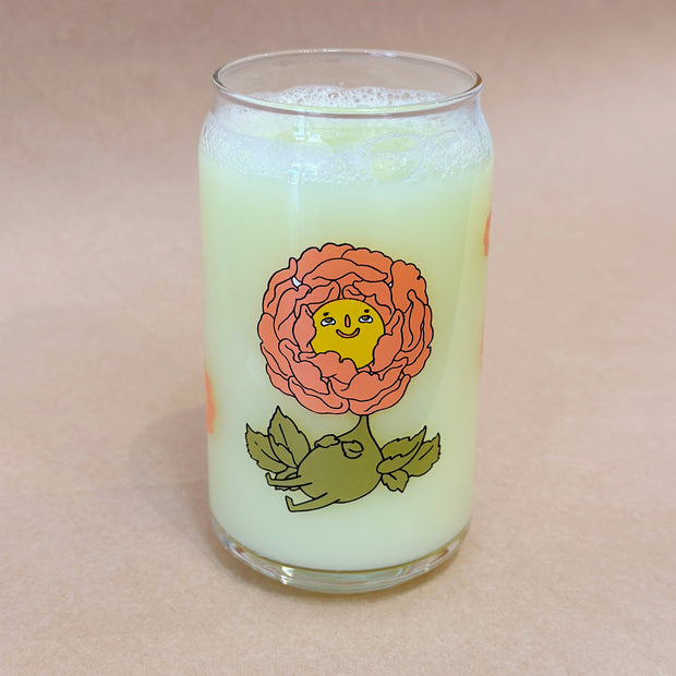 Glass cup with a flat base and slightly inward lip. Features a graphic of a cartoon style orange peony, sitting with a smiling face and a chubby green body made out of stems and leaves. Around the rest of the glass are orange flowers.