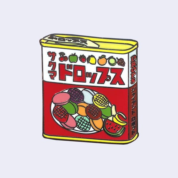 Die cut enamel pin of a tin of candy, Sakuma Drops, red and white packaging with drawings of fruit all over it. There is Japanese kanji written along the top and side of the container.