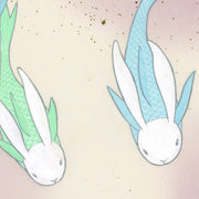 Paper circle with deckled edges and a soft olive green and cream colored watercolor background. Blue and green fish with white bunny heads swim calmly. Close up.