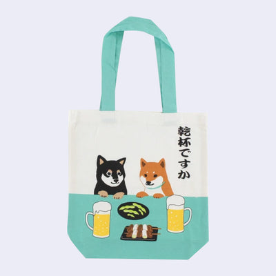 Cream colored tote bag with teal blue handle and color accents. Front features illustration of 2 shiba inus, sitting at a table with edamame, skewered food and 2 frothing beers.
