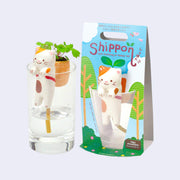 Ceramic calico cat, smiling and positioned over a small plastic cup. Its backing card features illustrations of plants and trees and reads "Shippon". 