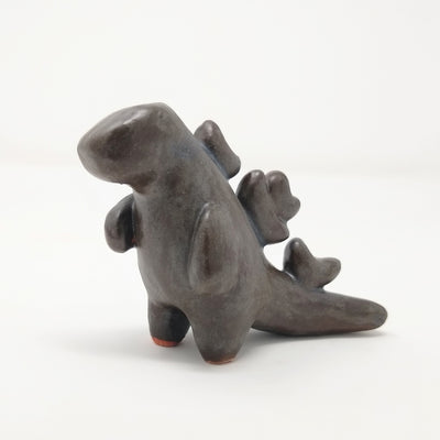 Dark brownish black ceramic sculpture of Godzilla, with minimalistic smooth features and shapes. It has stylized spikes running down its back and no facial features.