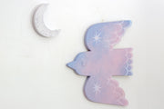 Die cut bird shaped canvas. A pink and blue painted bird flies with an eye closed and sparkles on its wings. A crescent moon hangs nearby.