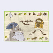 Polyproplyne mat, yellow background with a green outline featuring an illustration of Totoro playing a small wind instrument and holding an umbrella. Next to him, Catbus scurries away leaving paw prints. "My Neighbor Totoro" is written in the middle.