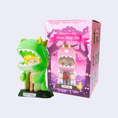 Small plastic figure of a blonde girl wearing a large dinosaur costume, designed to look like the Frog Prince. It is green with sad yellow frog eyes and a crown atop its head. It stands in front of its product packaging.