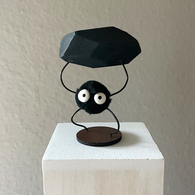 Small sculpture of a black soot sprite, with thin wiry arms and legs. It holds up a large piece of charcoal over its head.