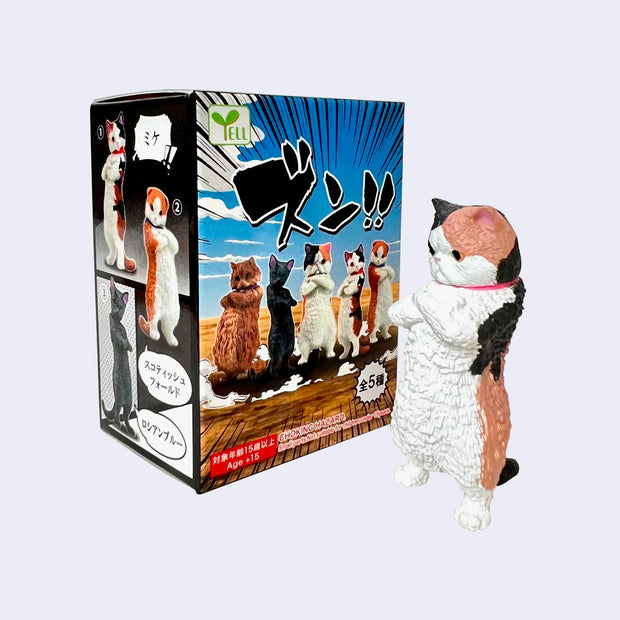Plastic figure of a chubby calico cat, standing on its hind legs with its arms crossed and an angry face. It stands next to its product packaging.