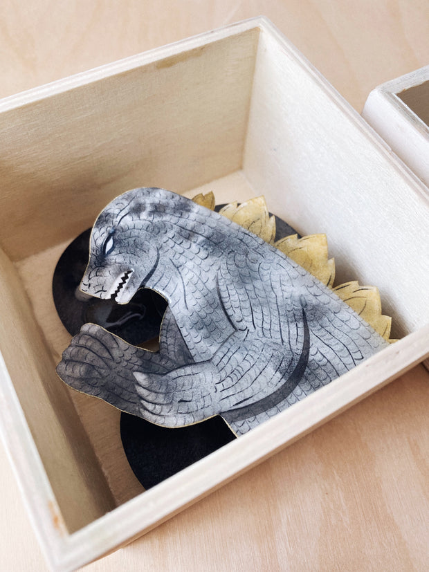 Side view of  a shadowbox-esque sculpture of Godzilla, colored gray with watercolor style pattern and gold spikes on its back. Only its upper body is showing.