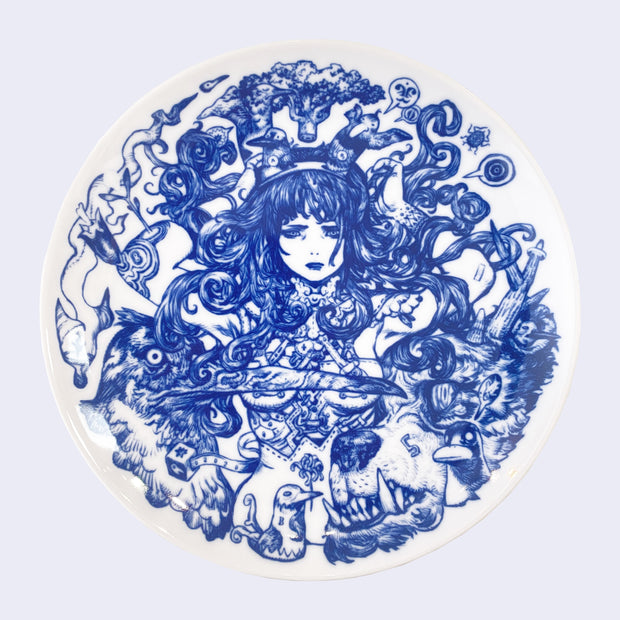 White ceramic plate with deep blue intricate line art. Plate features a woman with lots of dark, wavy hair, surrounded by a large bird with a long beak and a grizzled bear with an underbite.