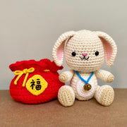 Crochet plush sculpture of a cute looking white rabbit with simple features and a smile, wearing a blue string with a bell on it. It is sitting next to a red pouch that is tied up with a gold string and has Chinese script on a gold patch on the center of the pouch.