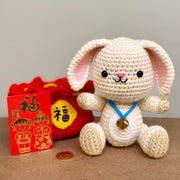Crochet plush sculpture of a cute looking white rabbit with simple features and a smile, wearing a blue string with a bell on it. It is sitting next to a red pouch that is tied up with a gold string and has Chinese script on a gold patch on the center of the pouch. Next to the rabbit is a red card with gold embossed New Years design.