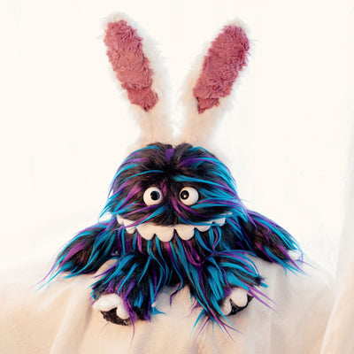 Plush doll of a monster creature, similar to a muppet, with a long underbite smile that wraps around its face and long black fur with blue and purple color highlights. It has wears a headband with white fluffy bunny ears on its head.