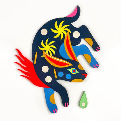 Flat wooden sculpture of a colorful blue rabbit, with its body turned to the side with polka dots and star patterns. A red flame comes out from below one of its arms and a green tear drop nearby.