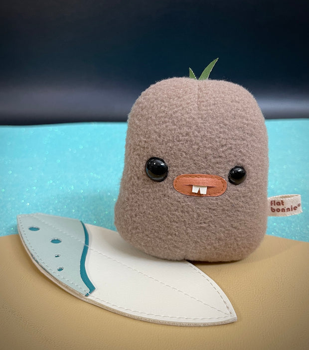 A brown small potato shaped plush with mismatched eyes, buckteeth and sprouts on its head. It has a small white and blue surfboard on the ground.